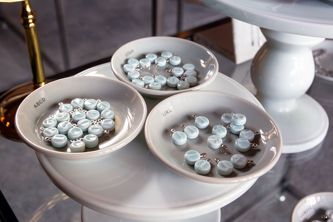 白い陶器の小皿に整列されたライトブルーの陶器製パーツとシルバー金具。各皿にアルファベット表記